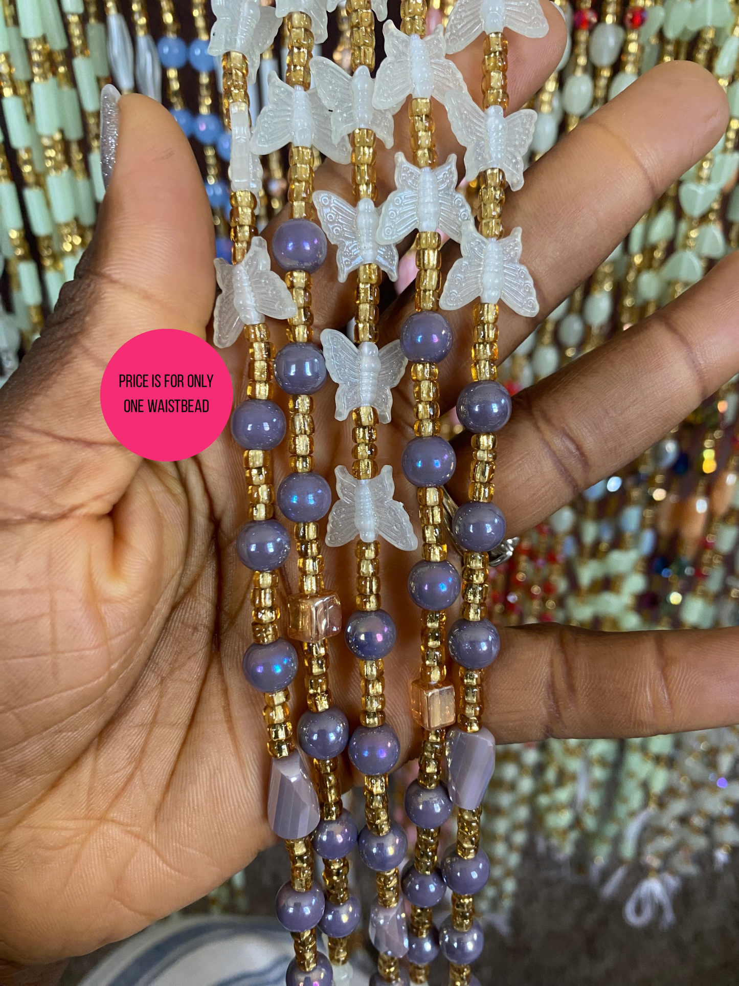 Hand holding a beaded waist bead with butterfly decorations, with a pink label on a blurred background.
